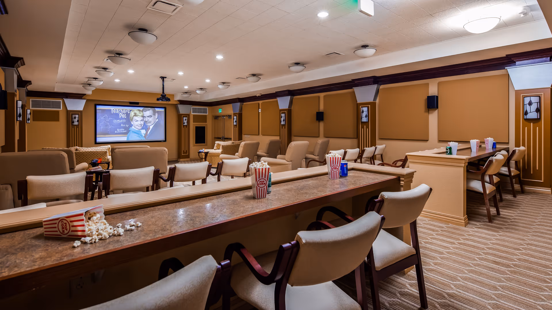 Interior view of a cozy movie theater room with multiple rows of beige cushioned chairs and a large screen displaying a black and white movie. Popcorn containers and soda cans are placed on the counter and tables, creating a relaxed and inviting atmosphere.