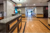 Reception desk and wide corridor of a nursing facility with wood-patterned floors, ceiling lights, and floral arrangement on the counter.