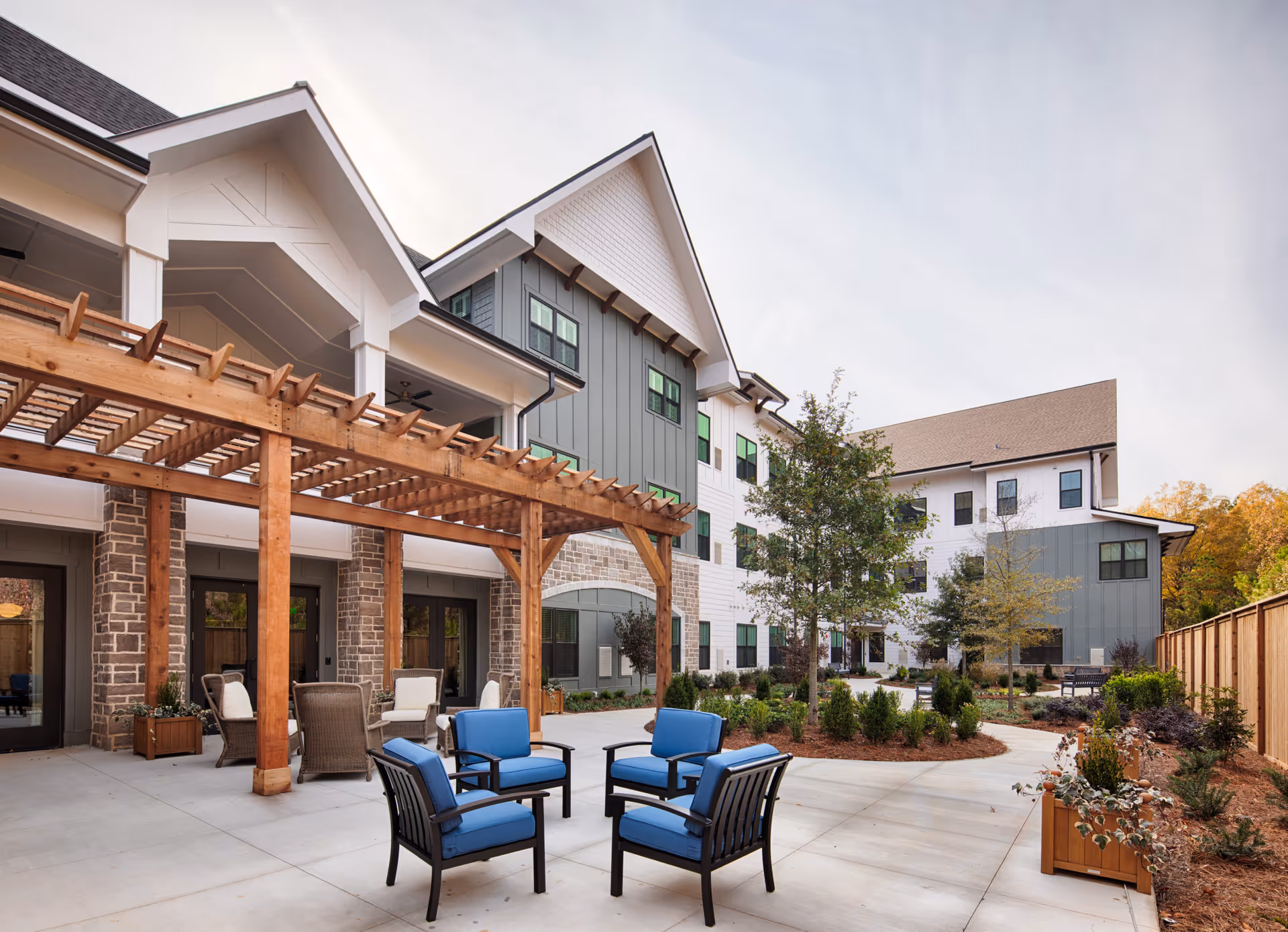 Outdoor patio area at Longleaf Liberty Park featuring a wooden pergola, several cushioned chairs arranged around a small table, landscaped garden beds, and a multi-story building with a mix of stone, white, and green siding in the background.