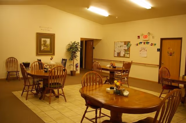 A communal dining room with multiple wooden tables and chairs, floral centerpieces, and a bulletin board on the wall.