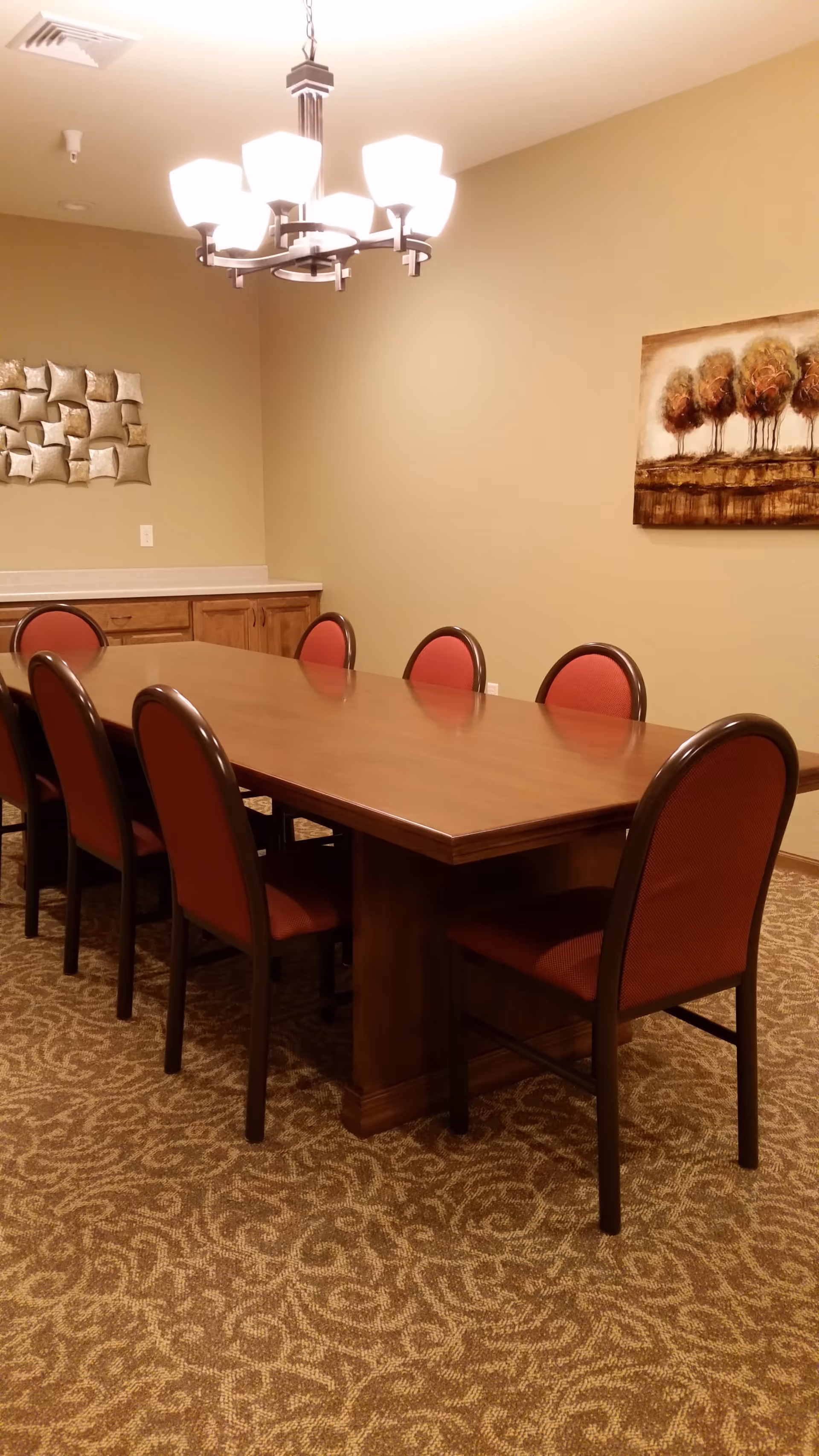 A dining room with a long wooden table surrounded by eight chairs with red upholstery. The room has beige walls, a patterned carpet, a modern chandelier with multiple lights hanging from the ceiling, and decorative wall art including a metallic abstract piece and a painting of trees.