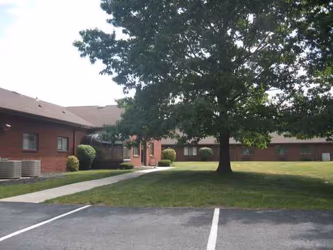 Brick senior living facility with a lawn, large tree, walkway, and parking.