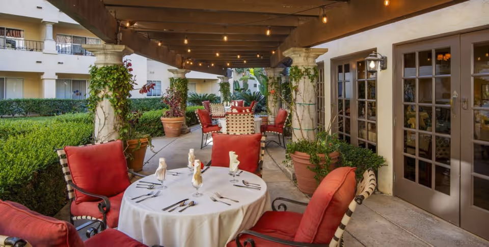 Outdoor covered patio area with round tables covered in white tablecloths, set with napkins and silverware. Red cushioned chairs surround the tables. Large potted plants and greenery decorate the space, with string lights hanging from wooden beams above. French doors lead inside the building.
