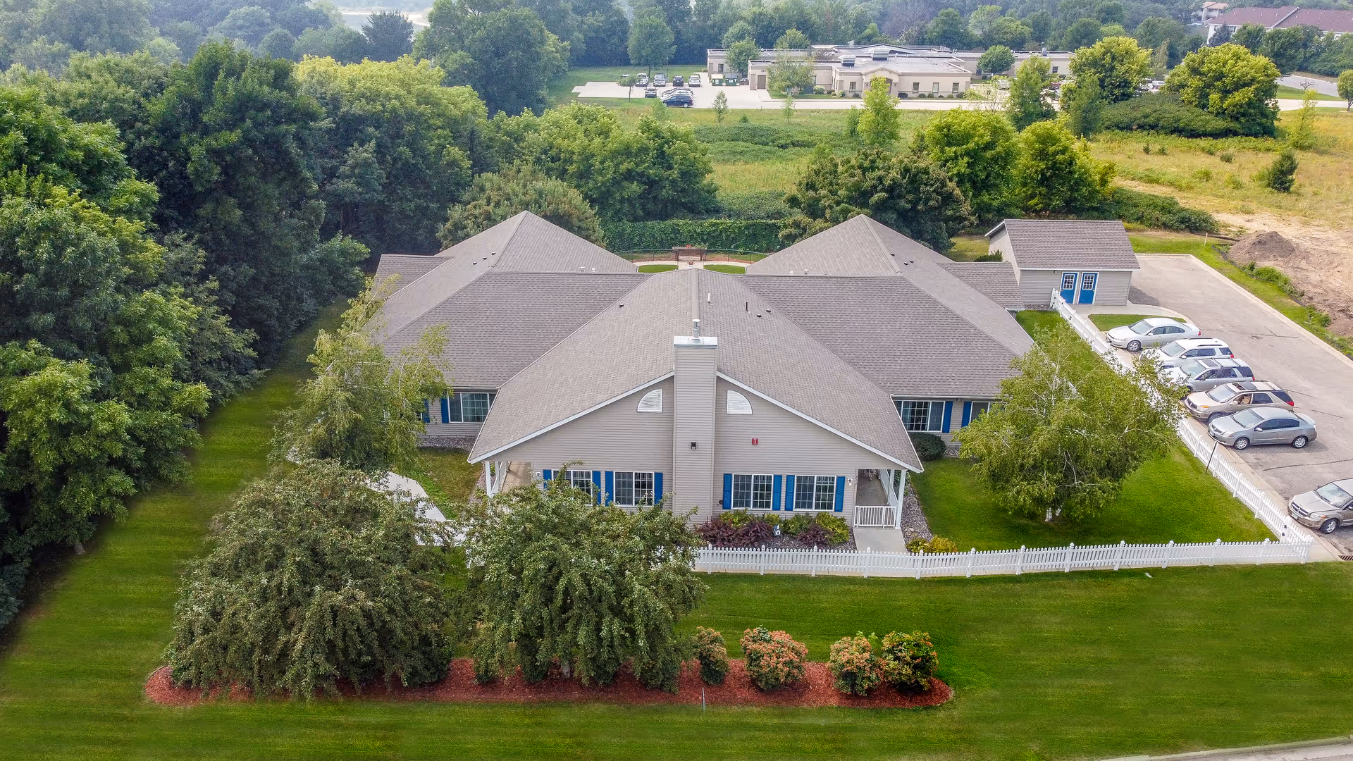Aerial view of a single-story residential building with a gray roof and beige siding, surrounded by green lawns, trees, and shrubs. There is a white picket fence enclosing part of the yard, and a parking lot with several cars is visible to the right. The building is set in a semi-rural area with other buildings and greenery in the background.