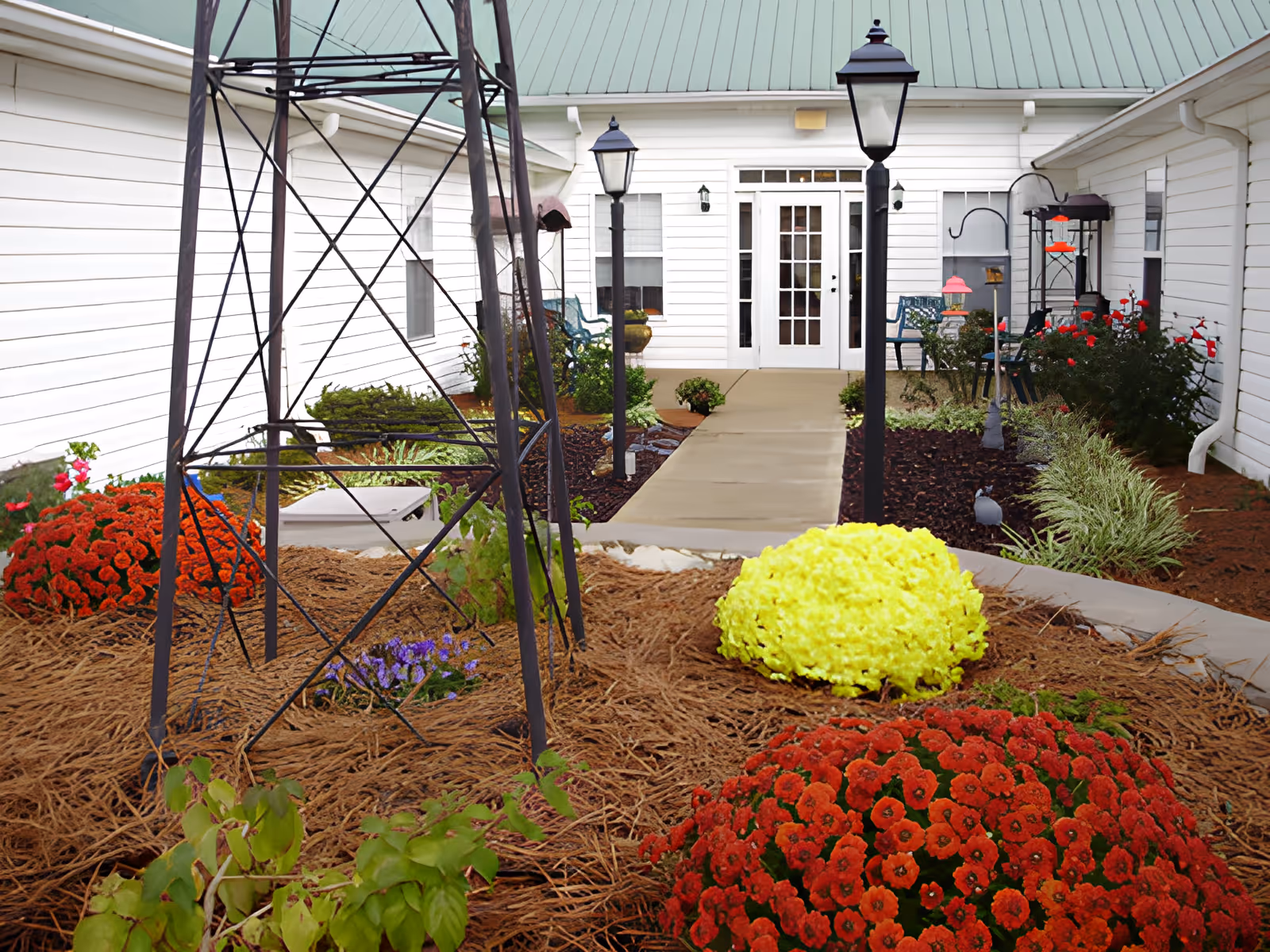 Courtyard garden with a paved walkway leading to a white building entrance, flanked by lampposts and colorful flowerbeds.
