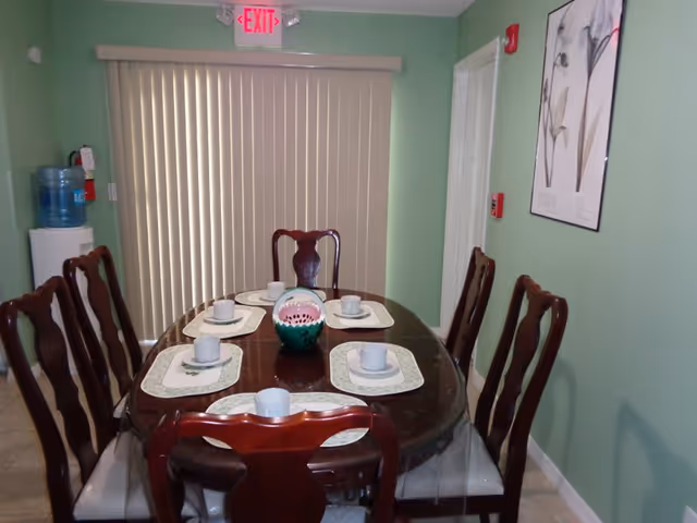 A dining room with a dark wooden oval table set with six place settings, each including a placemat, cup, and saucer. Six matching wooden chairs surround the table. A decorative vase is placed in the center of the table. The room has light green walls, a framed floral artwork on the right wall, vertical blinds covering a window or sliding door at the back, and a water dispenser with a fire extinguisher on the left side. An exit sign is visible above the blinds.