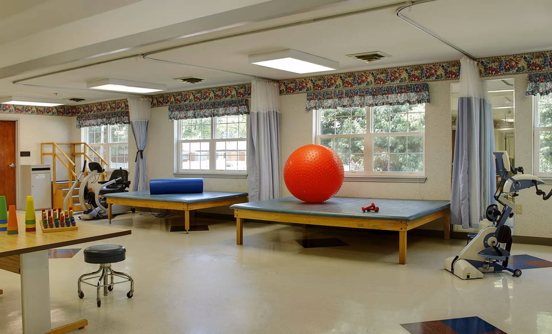 A bright therapy/exercise room with windows, raised treatment tables, exercise equipment and a large red therapy ball.