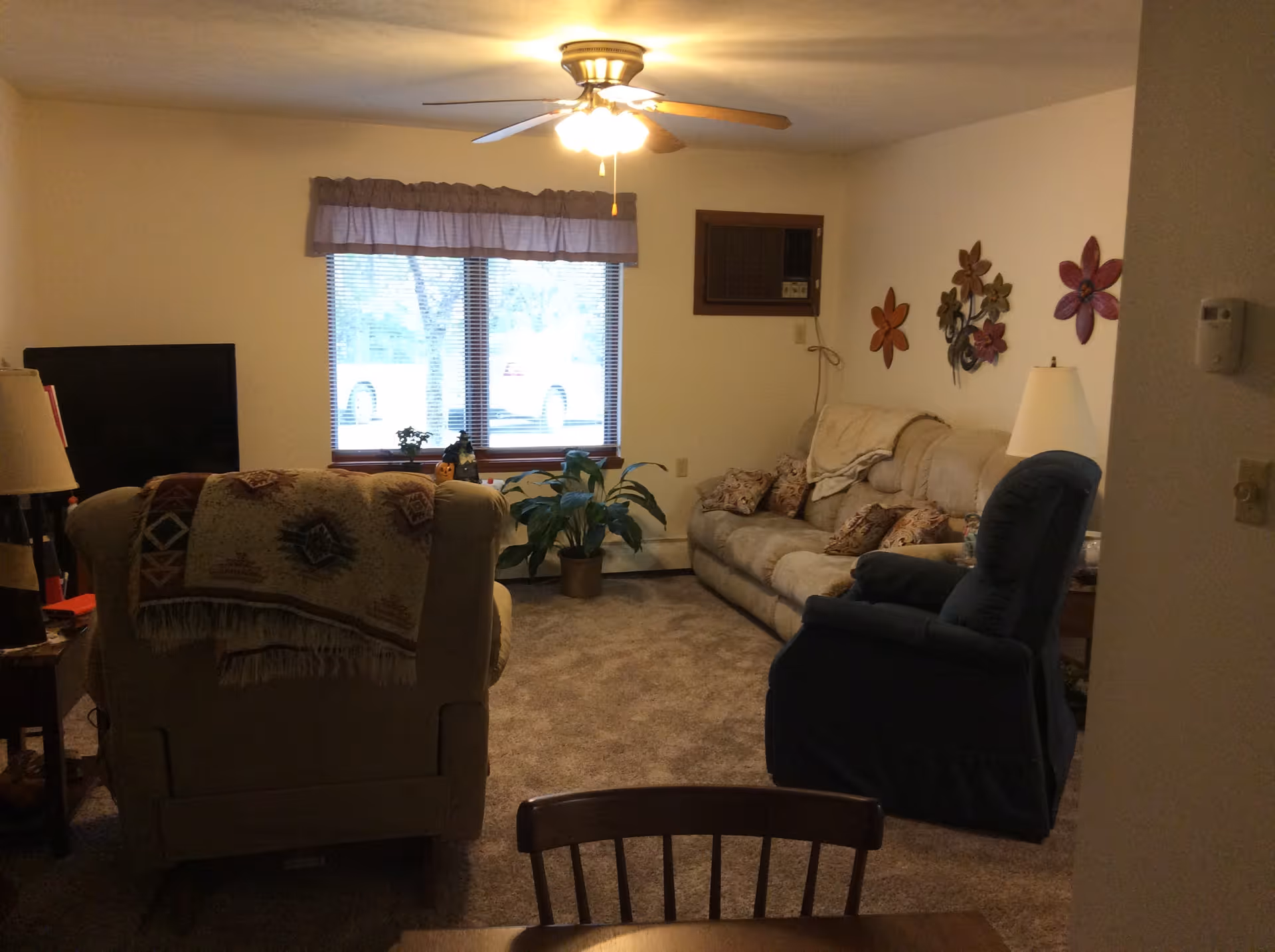 Cozy living room with beige carpet, a beige sofa with patterned pillows, a blue recliner, and a beige armchair with a patterned throw. A ceiling fan with lights is on the ceiling. There is a window with blinds and a valance, a potted plant near the window, and decorative flower wall art on the right wall. A TV is partially visible on the left side, and a table lamp is on a side table next to the recliner.