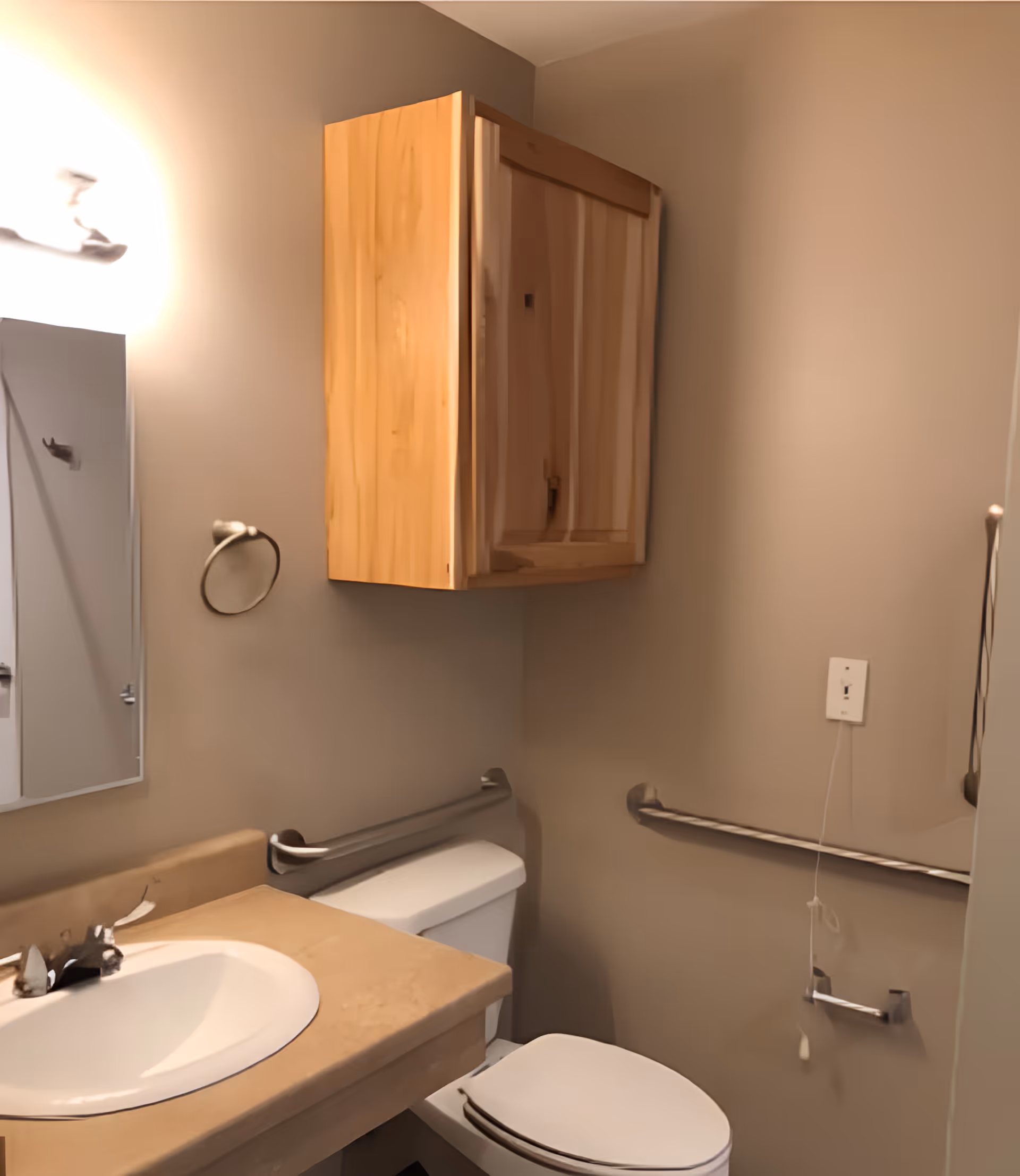 A small bathroom with a beige countertop sink, a white toilet, a wooden wall cabinet above the toilet, a mirror on the left wall, and metal grab bars installed on the walls near the toilet and sink. The walls are painted light beige.