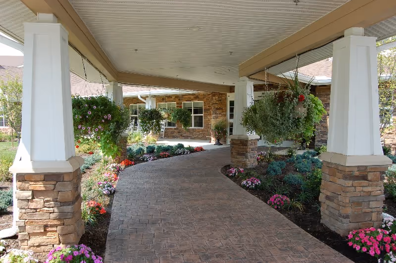 Covered walkway with stone pillars and hanging flower baskets leading to the entrance of a building surrounded by landscaped flower beds with various colorful flowers and greenery.