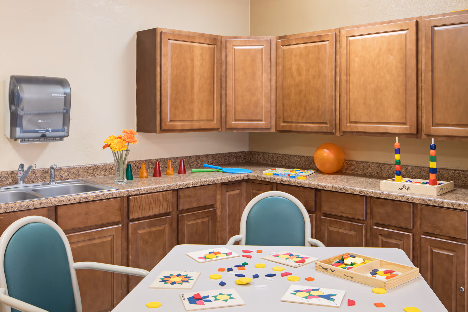 Small kitchen/activity room with wooden cabinets, a sink and a table covered in colorful therapy toys and puzzles.