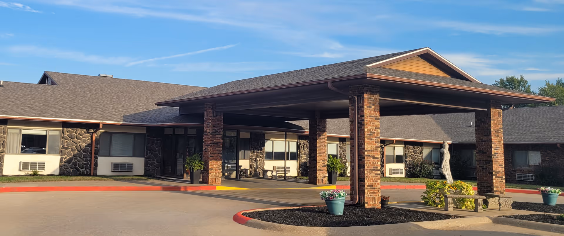 Front entrance of Belleview Care Center with a covered porte-cochere supported by brick columns, stone-faced building, potted plants, and a small statue.