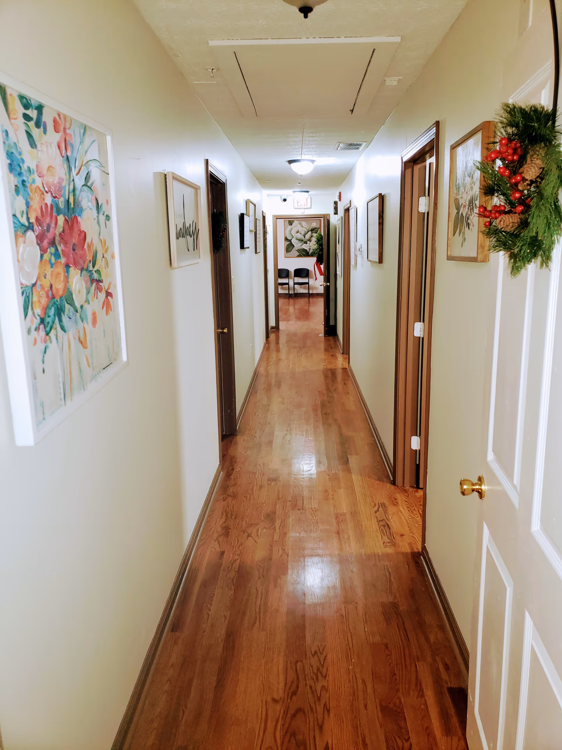 A long, narrow hallway with wooden flooring and cream-colored walls. Several doors line both sides of the hallway, some decorated with wreaths. The walls are adorned with framed floral artwork and decorative signs. At the end of the hallway, two black chairs are visible beneath a large floral painting. The ceiling has a light fixture and an access panel.