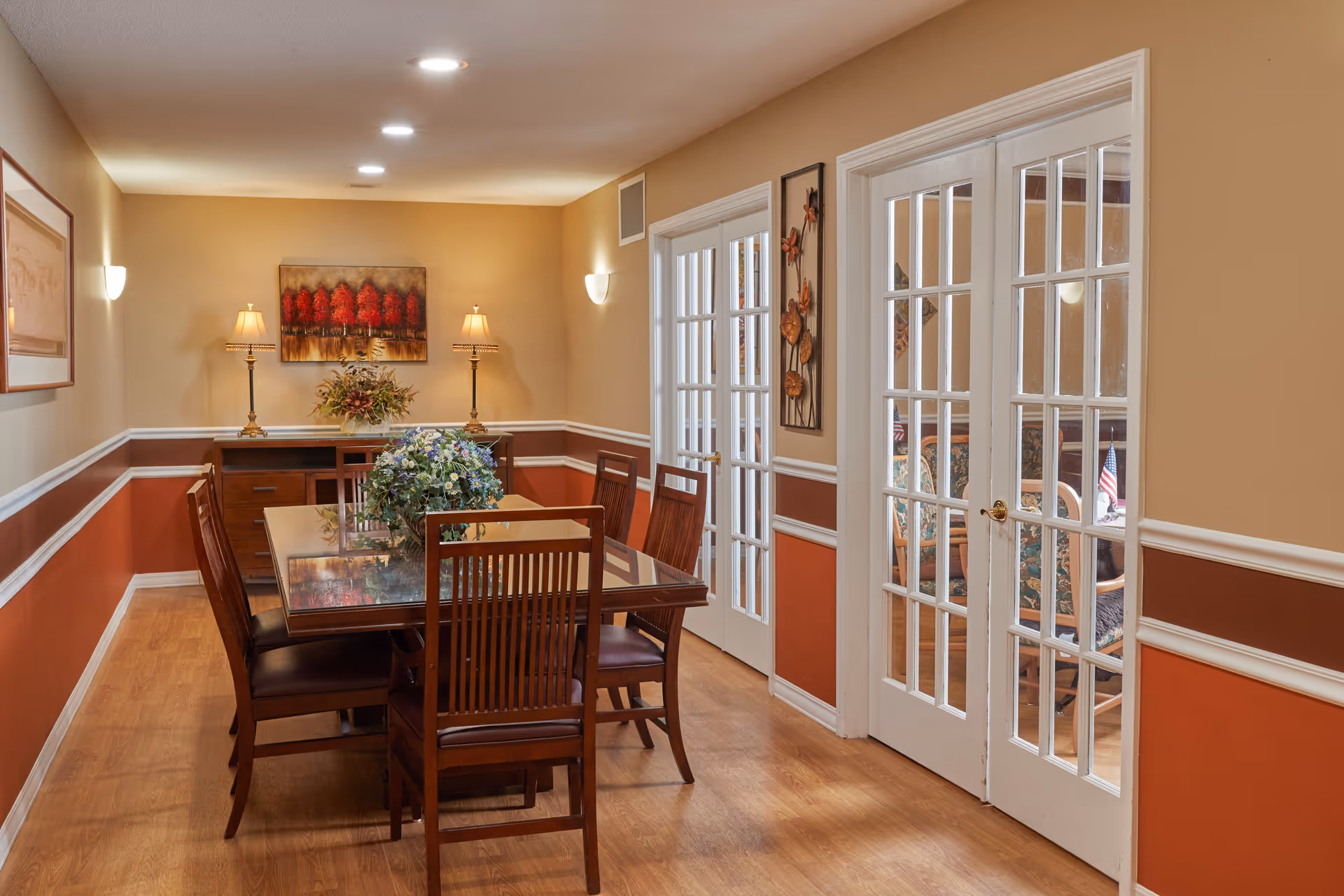 A dining room with a wooden table and six matching chairs. The table has a floral centerpiece. The walls are painted beige and rust with white trim. There are two table lamps on a sideboard against the far wall, and a painting of red trees hangs above it. On the right side, there are white French doors leading to another room with floral upholstered chairs.