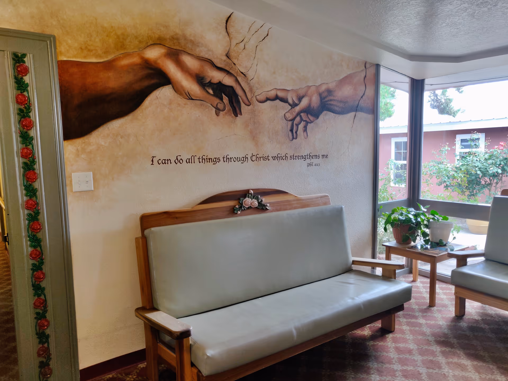 Seated lounge area with upholstered wooden benches beneath a mural of two hands reaching toward each other and windows looking out to a courtyard with plants.