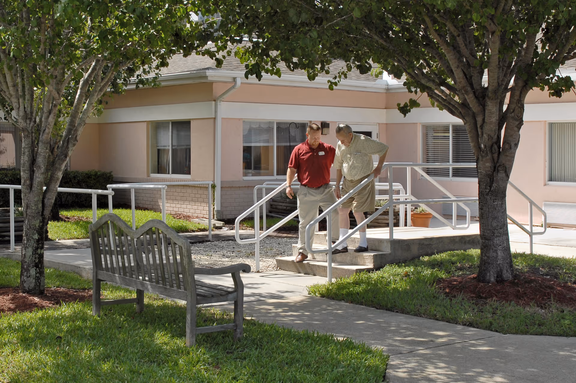 An outdoor scene at a senior living facility showing two men standing on a small set of concrete steps with metal handrails. One man, wearing a red shirt and beige pants, appears to be assisting the other man, who is wearing a light-colored shirt, shorts, and white socks. The area is landscaped with green grass, trees, and a wooden bench. The building in the background has light-colored walls and windows with blinds.