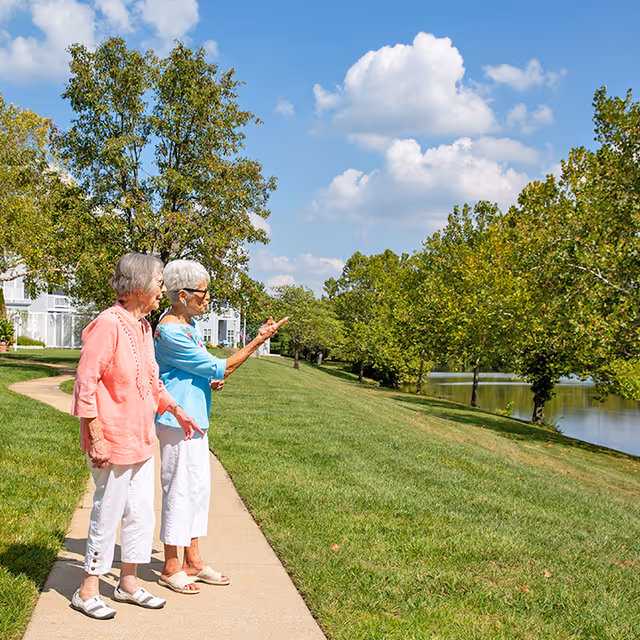 Two elderly women standing on a paved pathway in a grassy outdoor area near a body of water, surrounded by trees under a partly cloudy blue sky. One woman is wearing a coral top and white pants, and the other is wearing a light blue top and white pants, pointing towards something in the distance.
