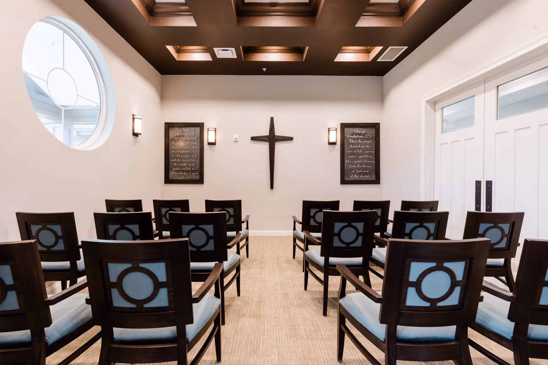 A small chapel room with rows of wooden chairs featuring blue cushions and decorative backs. The room has a beige carpet, white walls, and a dark coffered ceiling. On the front wall, there is a wooden cross flanked by two framed scripture plaques and wall sconces. A large circular window is visible on the left side, and double white doors are on the right.