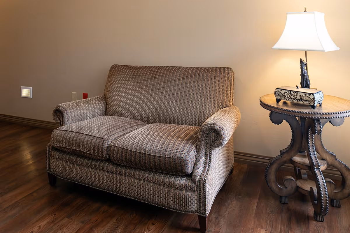 A patterned loveseat sofa next to a wooden side table with a decorative box and a lit table lamp, set against a beige wall and wooden floor.