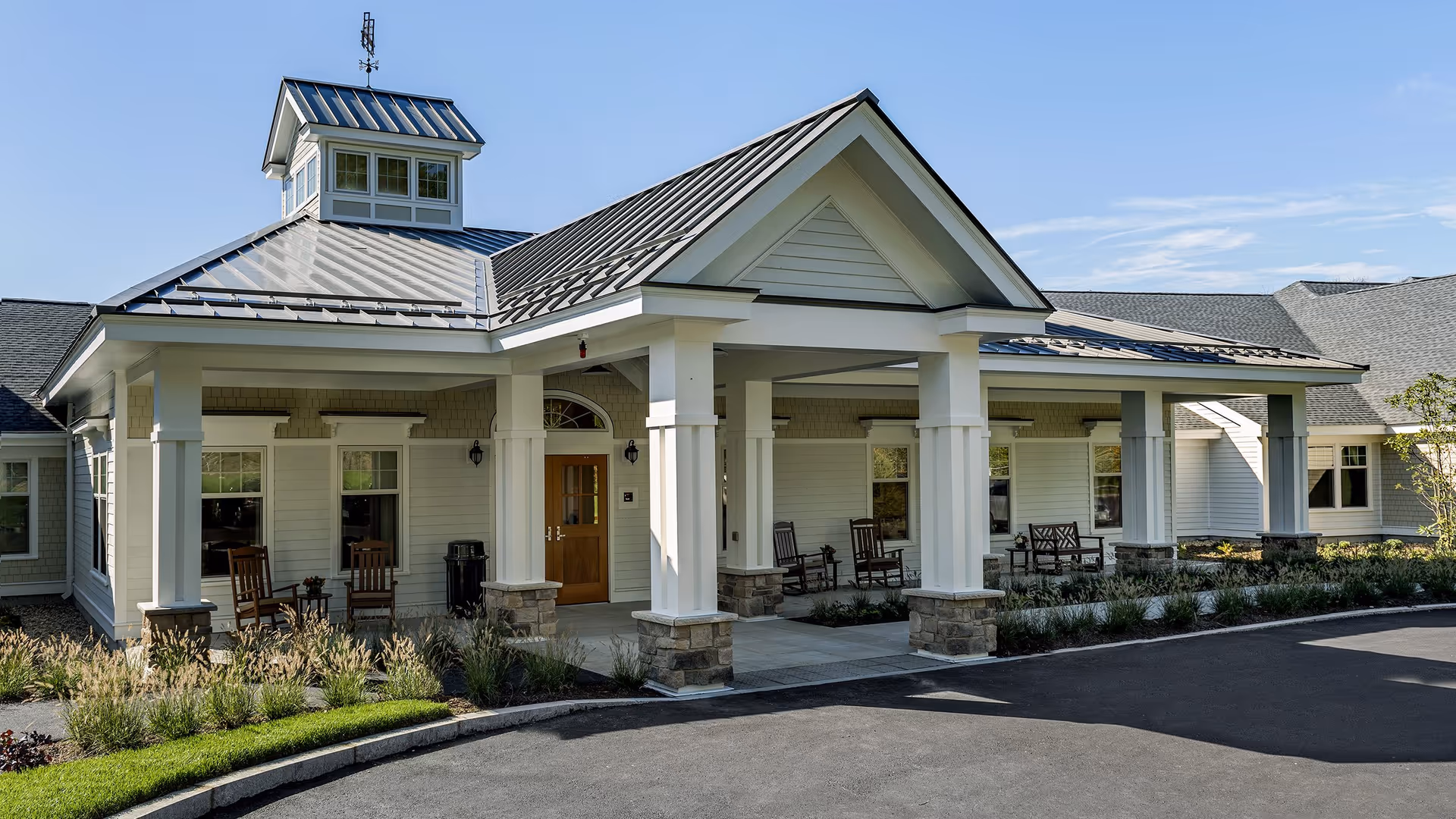 Front entrance of a single-story residential facility with a covered portico, white columns, rocking chairs, and a paved driveway.