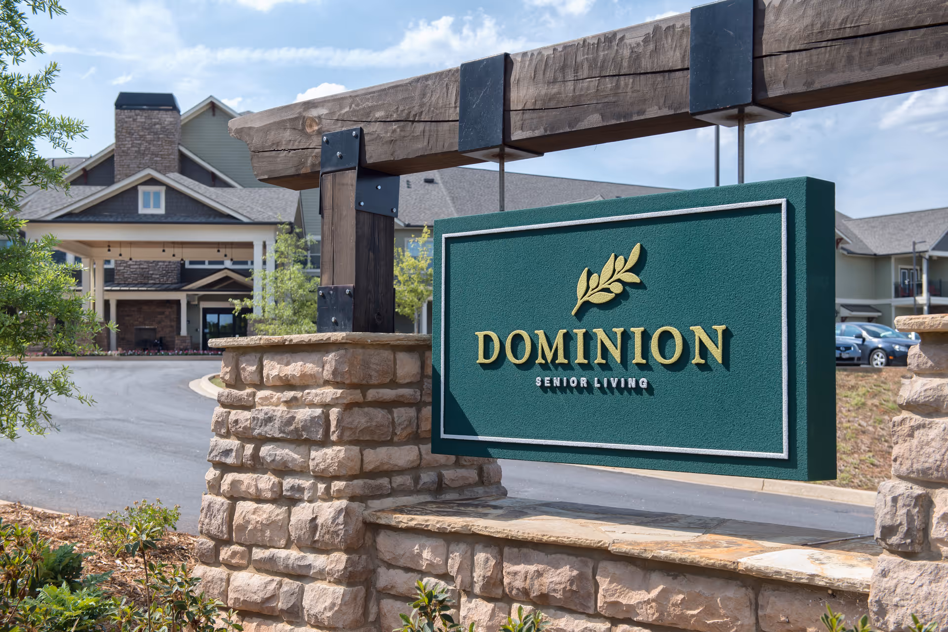 Stone and wood entrance sign for Dominion Senior Living with the senior living facility building visible in the background under a partly cloudy sky.