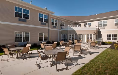 Outdoor patio area of a senior living facility with multiple round tables and chairs arranged on a concrete surface, surrounded by a two-story beige building with many windows under a clear blue sky.
