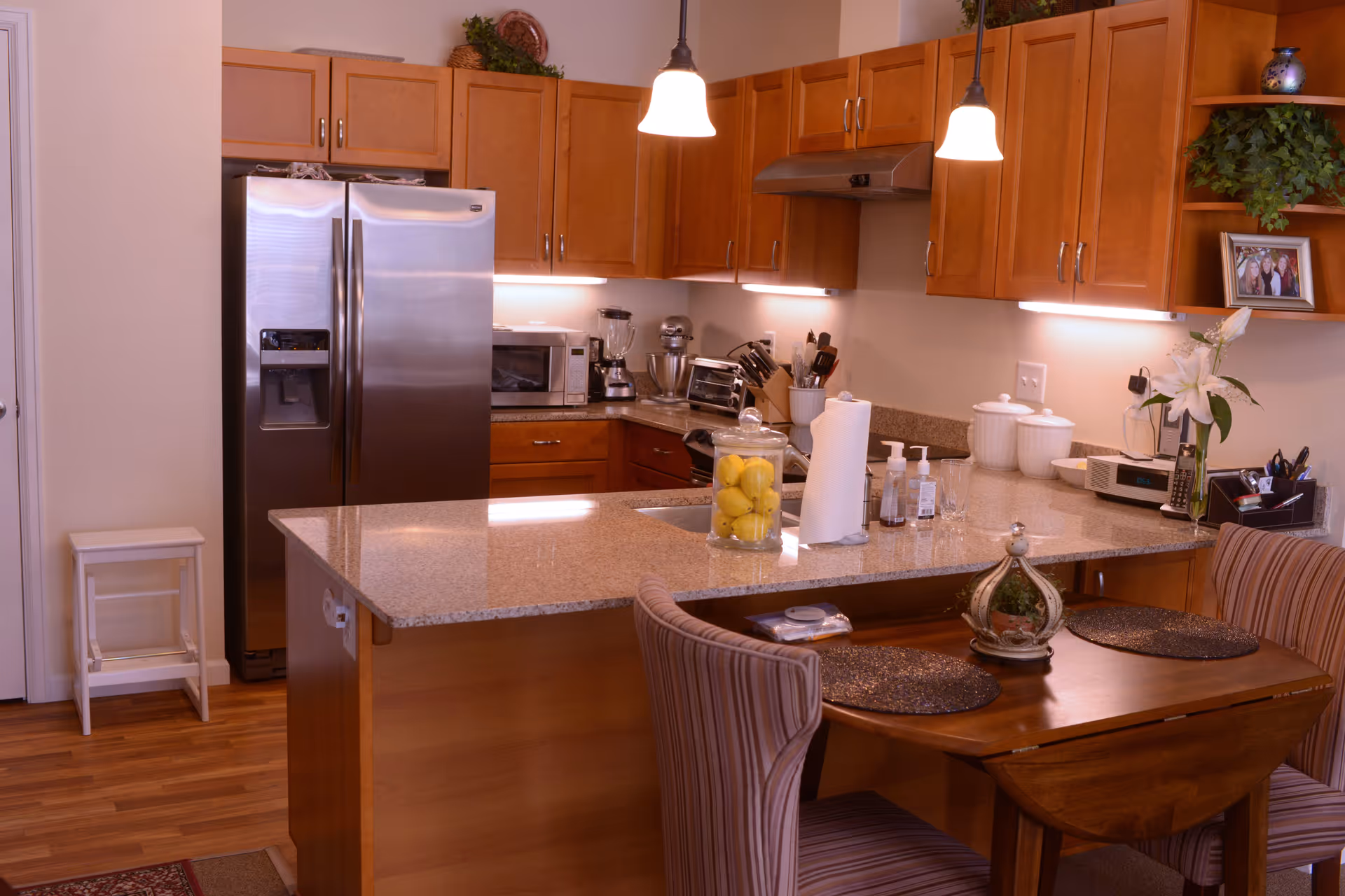 A modern kitchen with wooden cabinets, a stainless steel refrigerator, a microwave, a blender, and various kitchen utensils on the countertop. There is a granite island with a glass jar filled with lemons, paper towels, and hand soap. Two pendant lights hang above the island, and a small dining table with two striped chairs is adjacent to the island. The kitchen has wooden flooring and some decorative plants and framed photos on the shelves.