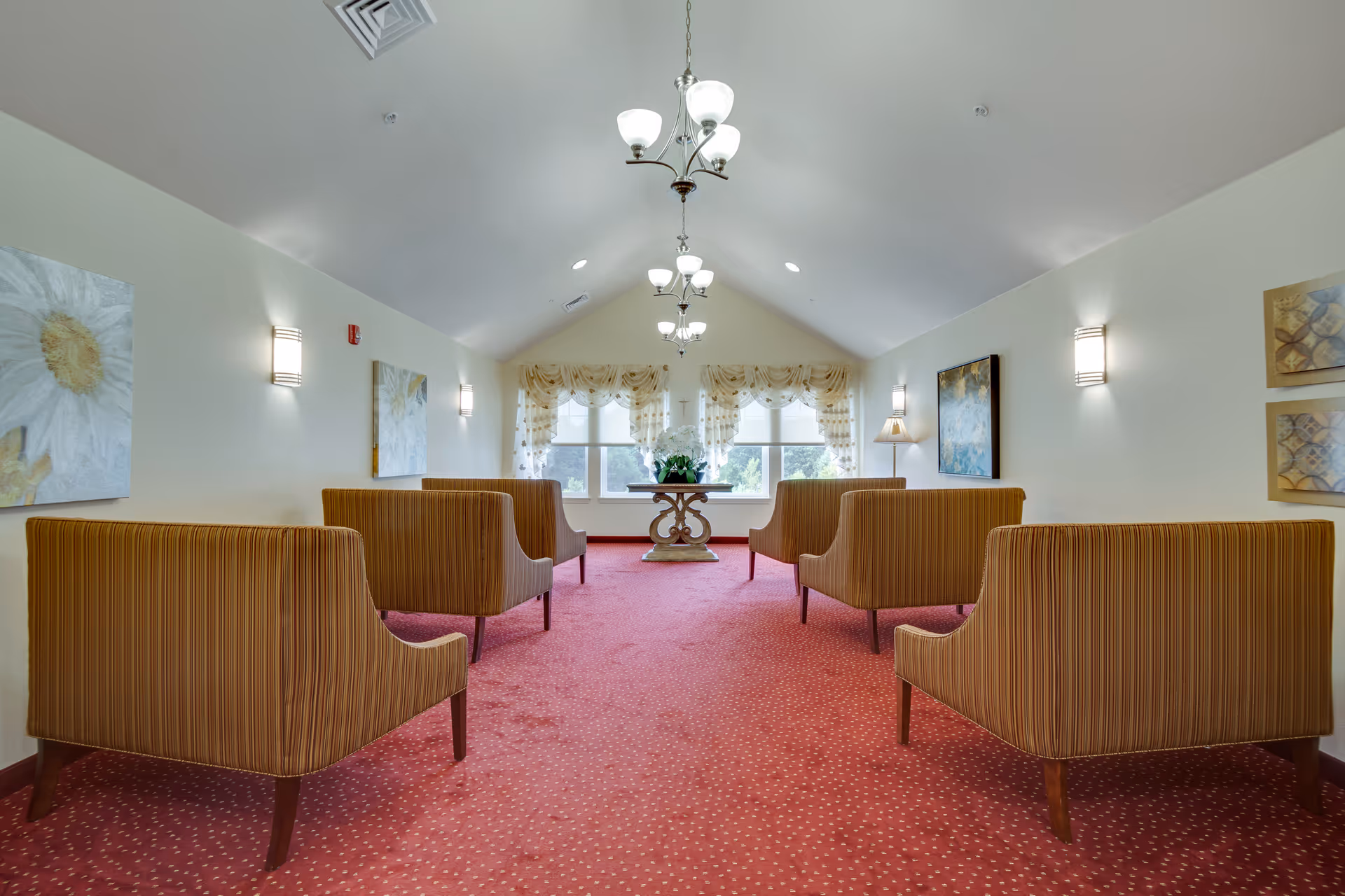 A bright, carpeted room with six striped armchairs arranged in two rows facing a window with sheer curtains. A decorative table with a flower arrangement is positioned in front of the window. The walls are adorned with paintings and wall sconces, and three chandeliers hang from the vaulted ceiling.