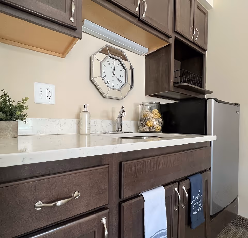 A kitchenette area with dark wooden cabinets, a white marble countertop, a small sink with a chrome faucet, a soap dispenser, a decorative glass jar filled with woven balls, a small refrigerator, and a wall clock with Roman numerals. Two towels hang from the cabinet handles, one white and one dark blue with the text 'wash your hands'.