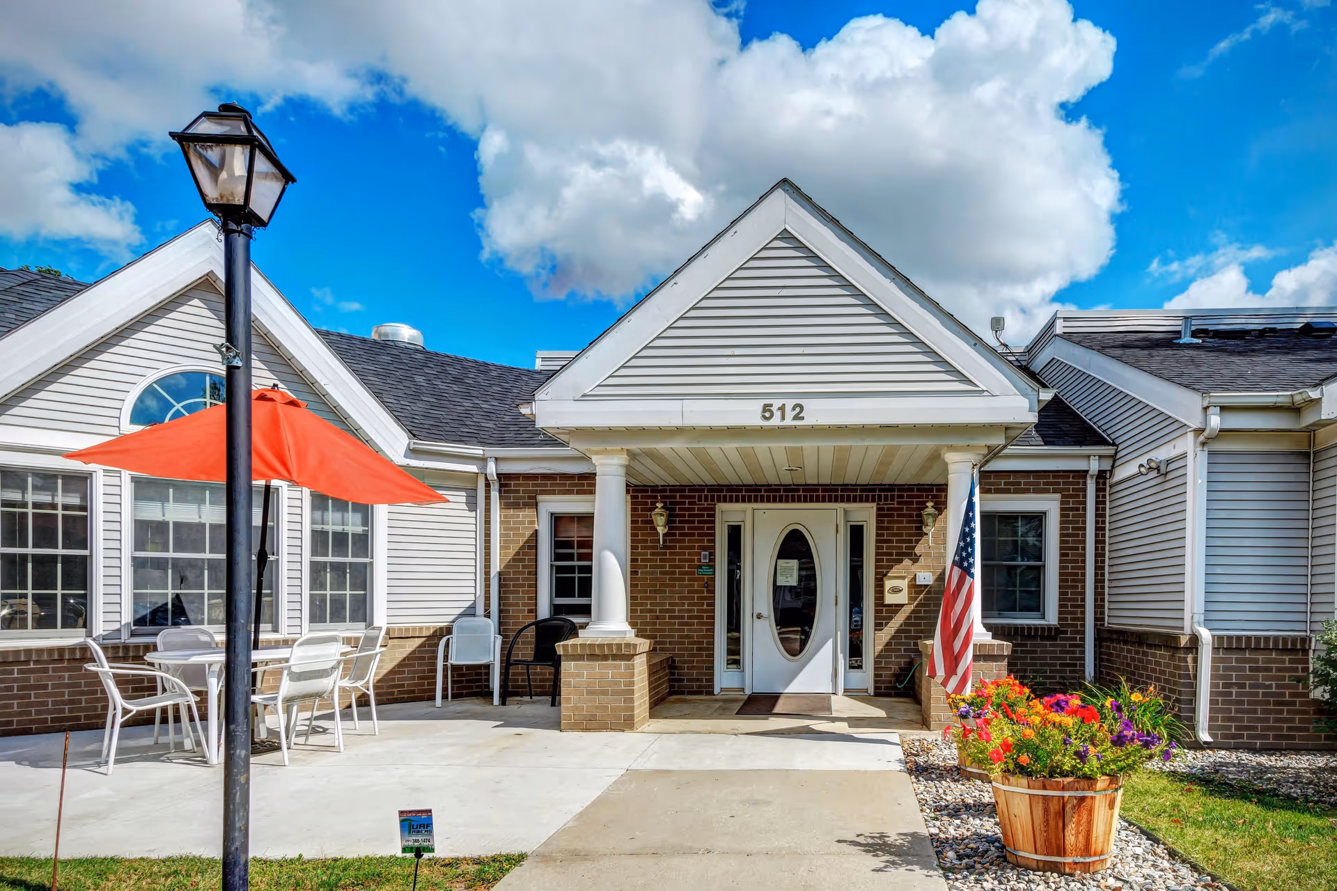 Front entrance of a senior living facility with the address number 512 above the door. The building has a brick and siding exterior with a covered porch supported by white columns. There is an American flag and a large wooden planter with colorful flowers on the right side. On the left side, there is a patio area with white chairs and a table under a red umbrella. A black lamp post is visible in the foreground. The sky is blue with scattered clouds.