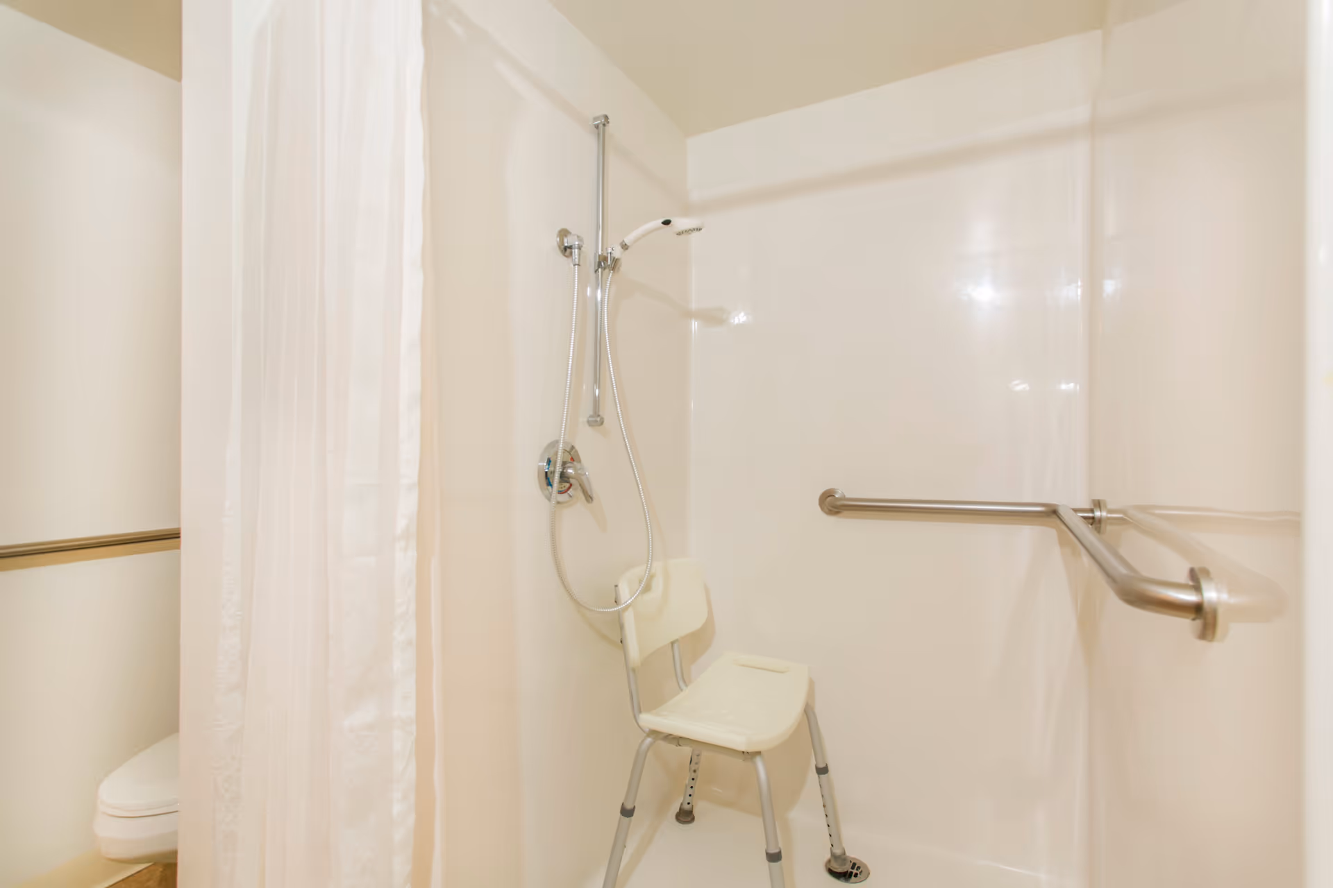 A shower area with a white shower chair, a handheld showerhead attached to a vertical bar, and a horizontal grab bar on the wall. A white shower curtain is partially visible, and a toilet can be seen in the background.