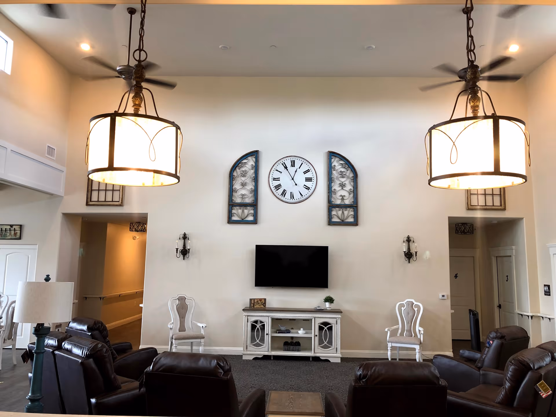A spacious living room area with high ceilings featuring two large hanging light fixtures and ceiling fans. The room has a wall-mounted flat-screen TV above a white console table, flanked by two decorative chairs. Several leather recliners are arranged facing the TV. The walls are light-colored with decorative wall art and a large clock above the TV. There are doorways on either side of the TV leading to other rooms.