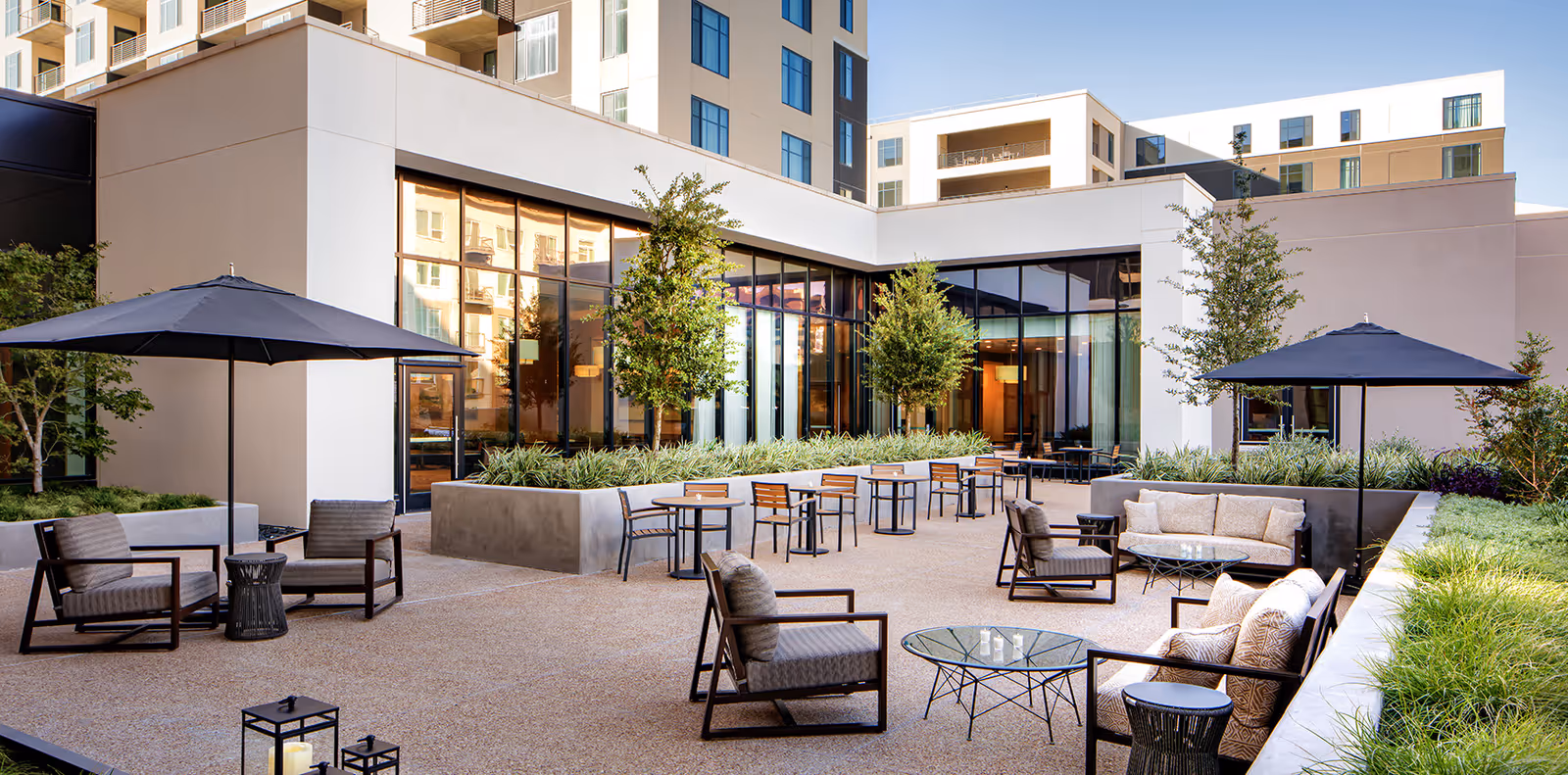 Outdoor patio area with modern seating including cushioned chairs, sofas, glass-top tables, and large black umbrellas. The space is surrounded by planters with greenery and small trees, adjacent to a building with large windows reflecting the surroundings.