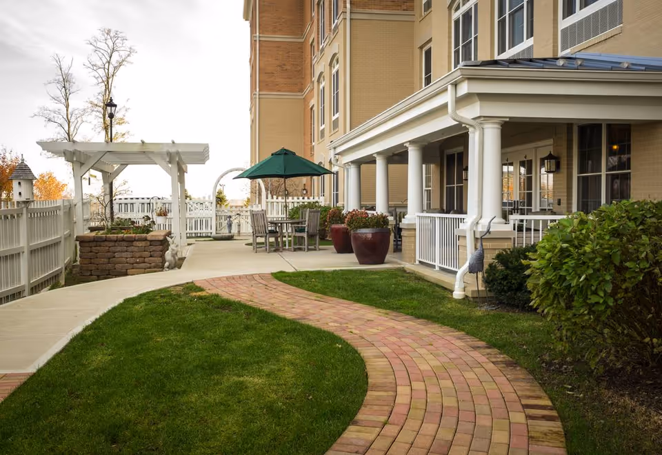 Outdoor patio area at Sunrise on Old Meridian featuring a curved brick pathway through green grass, a white pergola, a white picket fence, patio furniture with a green umbrella, large potted plants, and a beige building with white columns and windows.