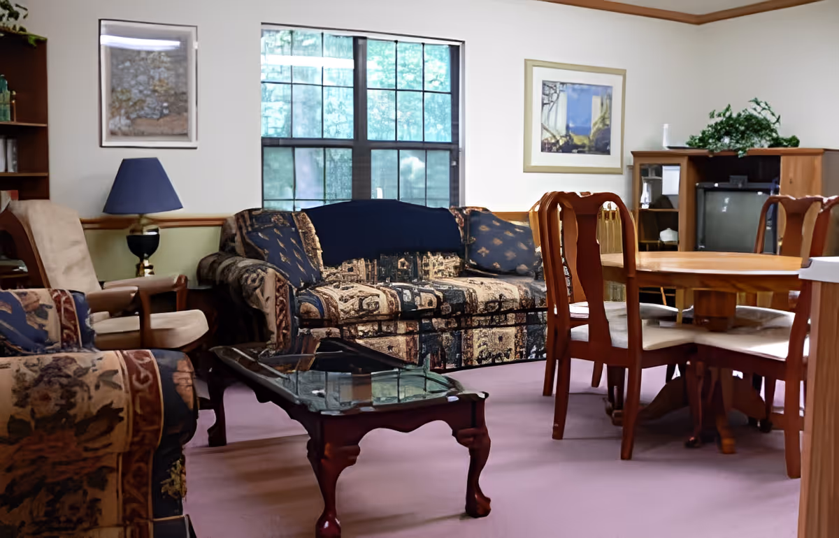 Cozy common room with patterned sofa and armchairs, glass-top coffee table, wooden dining table and chairs, and a TV cabinet by a window.