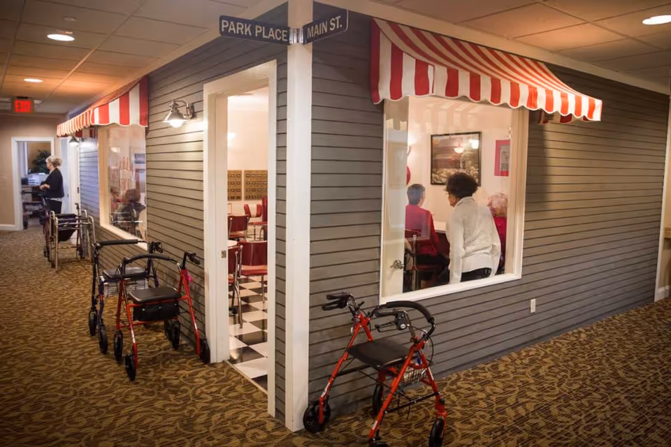 Indoor hallway of an assisted living facility with faux storefront facades, several red walkers parked along the wall, and residents visible through a window.