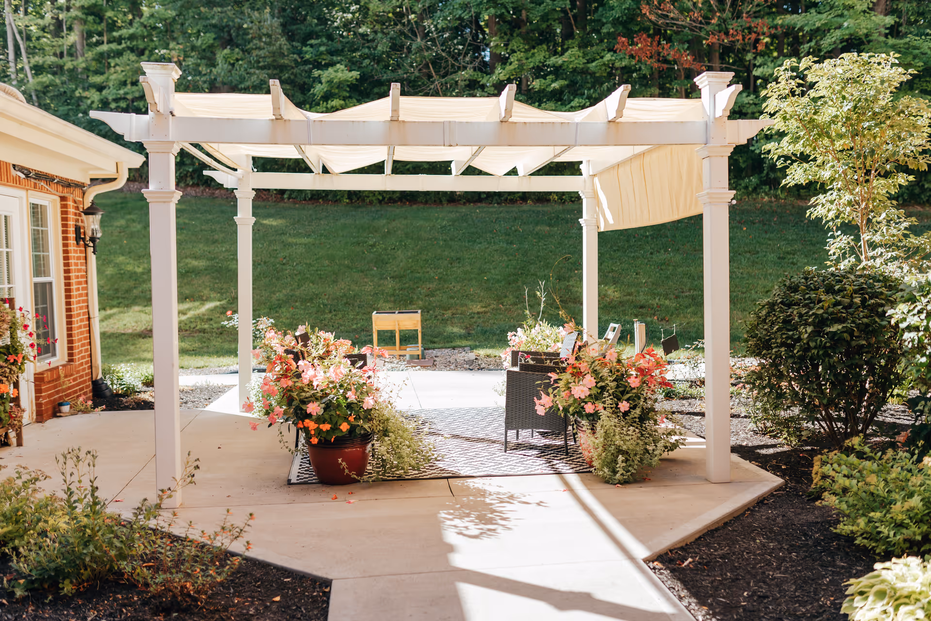 Outdoor patio area with a white pergola providing partial shade over a seating area with chairs and a small table. The patio is decorated with large pots of colorful flowers and surrounded by green bushes and trees. A brick building is visible on the left side.