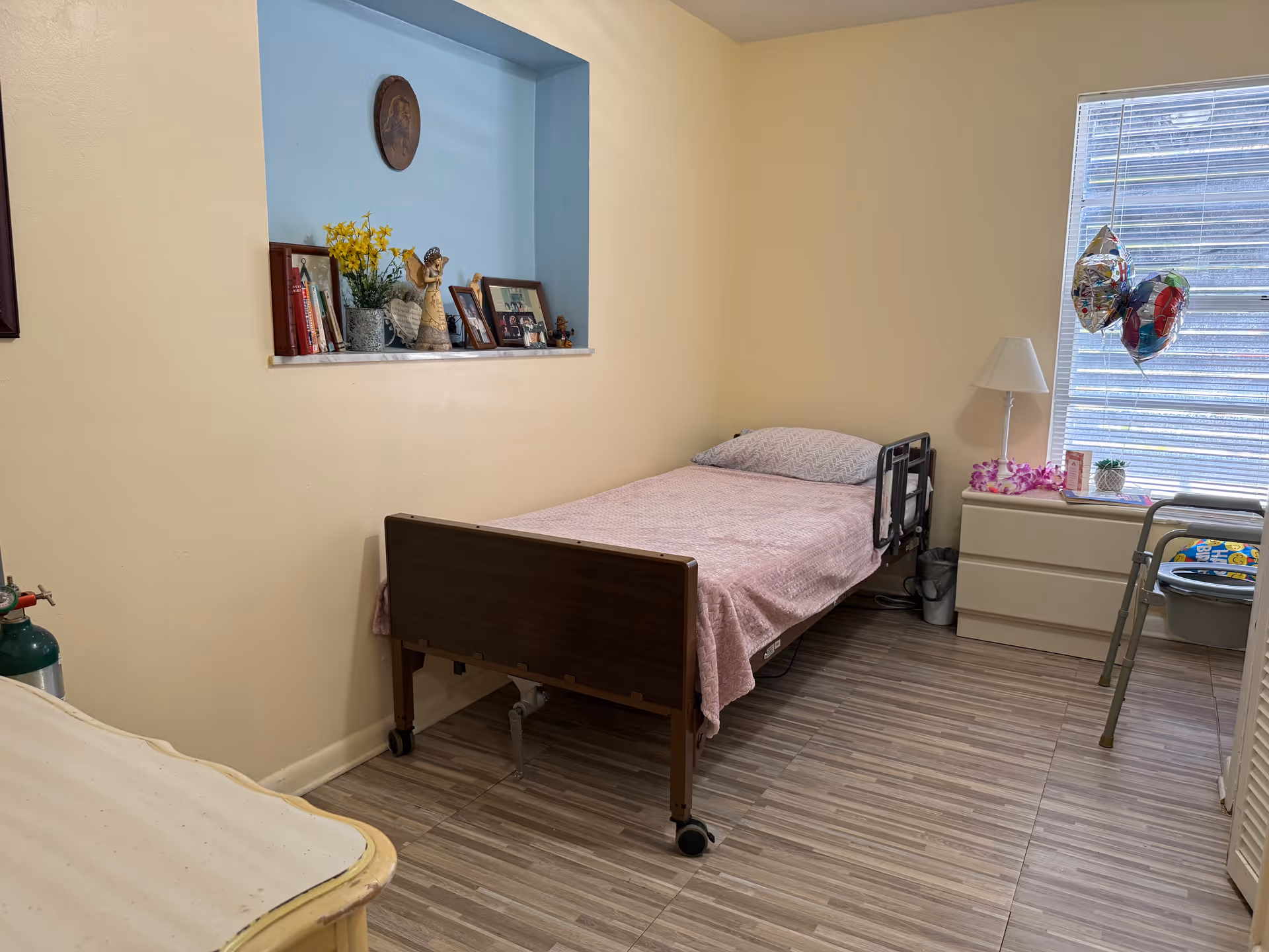 A small bedroom with a single hospital-style bed covered with a pink blanket and a pillow. There is a window with blinds partially open, a white dresser with a lamp, balloons, and small decorative items. A walker is positioned near the dresser. The walls are light yellow with a blue recessed shelf holding books, a vase with yellow flowers, framed photos, and decorative figurines. The floor has wood-patterned tiles.