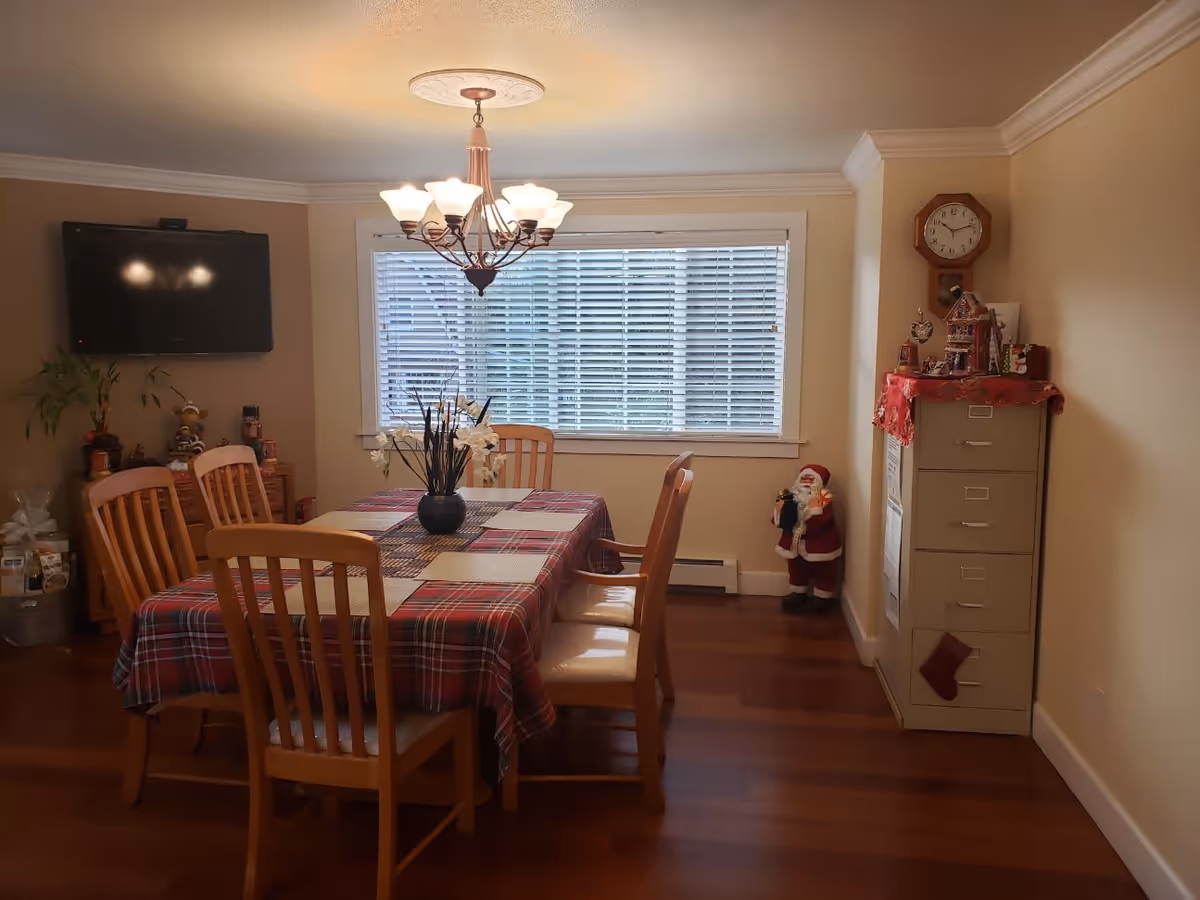 Dining room with a plaid-covered wooden table and chairs under a chandelier, window blinds, wall-mounted TV and a filing cabinet with holiday decorations.