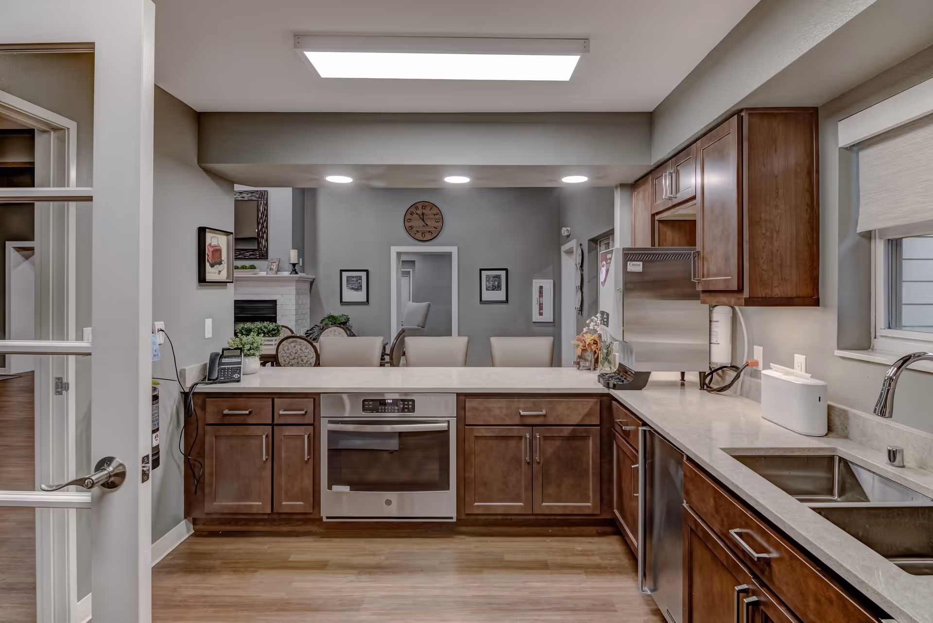 Interior view of a modern kitchen with wooden cabinets, a built-in oven, a dishwasher, and a double sink. The kitchen opens to a dining area with chairs and a wall clock above a doorway. The room has a bright ceiling light and neutral-colored walls.
