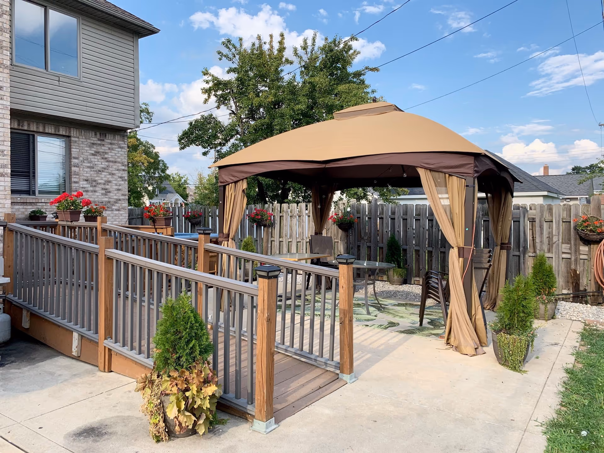 Outdoor patio with a tan gazebo, wooden ramped deck, potted plants, and a fenced yard beside a brick house.
