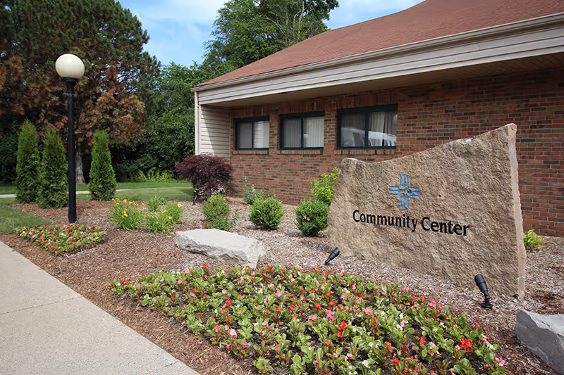 Exterior view of a community center building with a brick facade, surrounded by landscaped flower beds, shrubs, and a lamppost along a sidewalk.