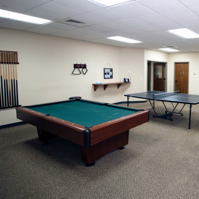 A recreational room with a green felt pool table and a ping pong table. The room has beige walls, a carpeted floor, and a ceiling with fluorescent lights. Pool cues are mounted on the wall, and a small shelf with a few items is also visible.
