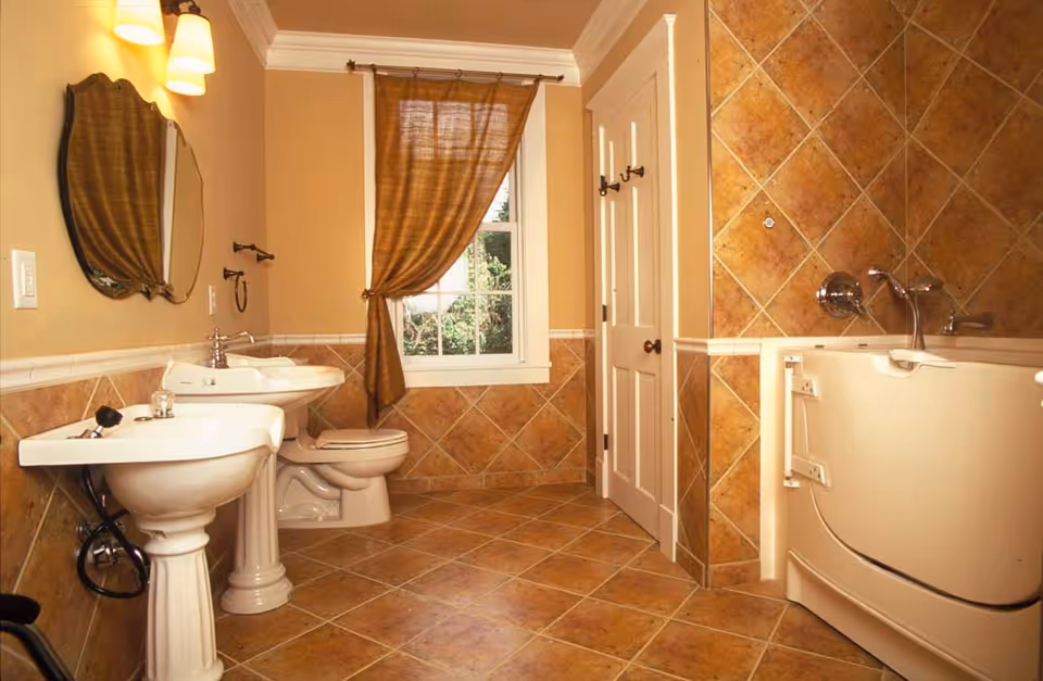 A bathroom with beige and brown tiled walls and floor, featuring a walk-in bathtub with safety handles, a toilet, two pedestal sinks with a mirror above one, a window with a brown curtain, and a white door with hooks.