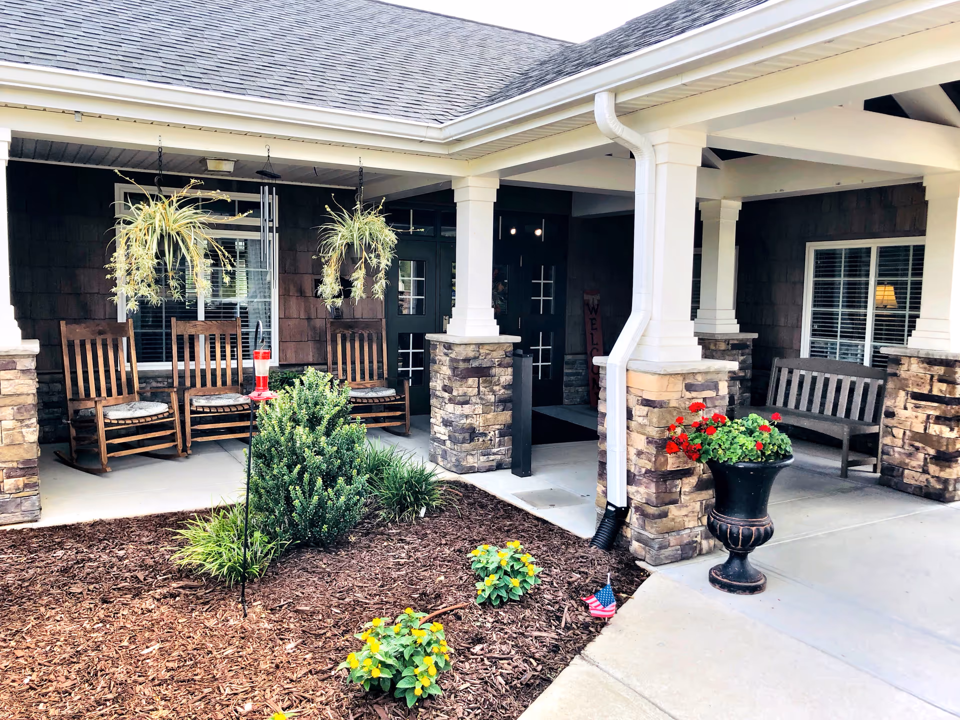 Covered porch area of Gabriel Manor featuring wooden rocking chairs, hanging plants, a small garden with flowers and shrubs, stone pillars, and a bench near the entrance door.