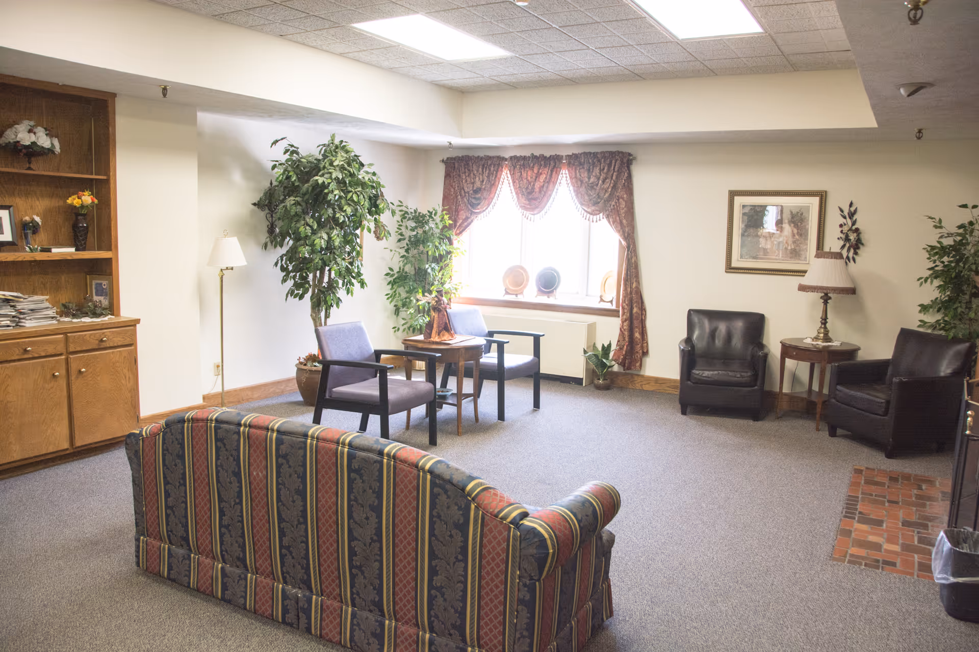 A cozy living room area with a striped sofa in the foreground, two armchairs with a small table between them near a window with patterned curtains, two black leather chairs with a side table and lamp, potted plants, a wooden cabinet with shelves, and carpeted flooring.