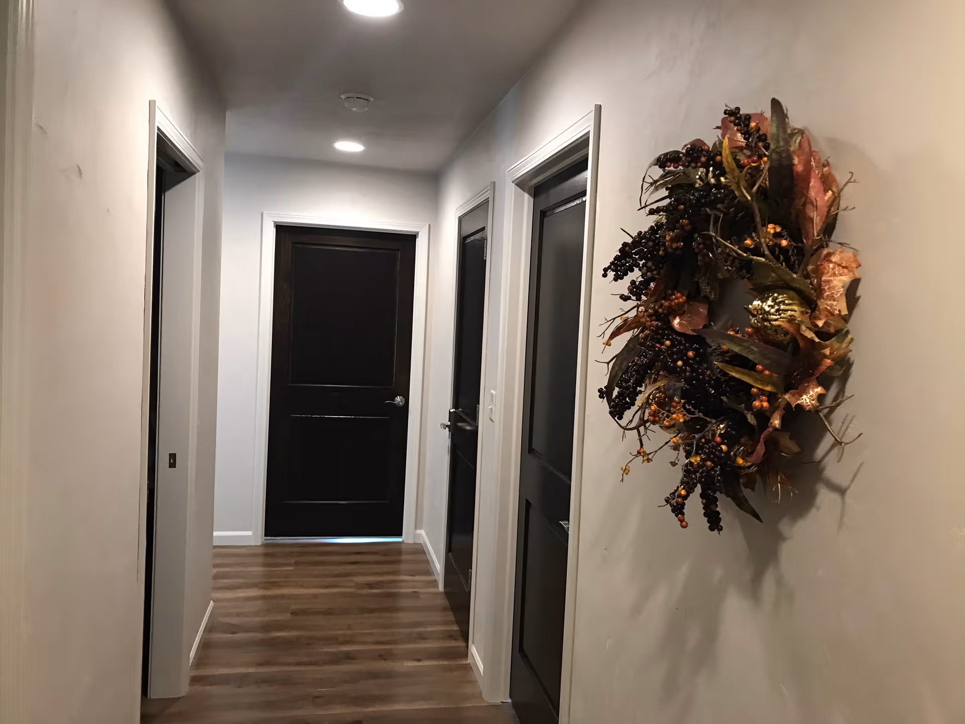 Interior hallway with wood-look flooring, dark doors, recessed ceiling lights, and a decorative wreath on the right wall.