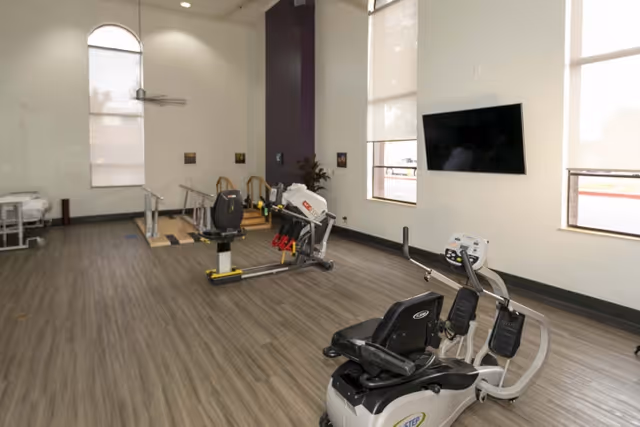 Bright therapy/exercise room with recumbent exercise machines, parallel bars, and a wall-mounted TV.