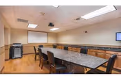 A conference or meeting room with a long rectangular table surrounded by black chairs. The room has wood flooring, beige walls with a chair rail, and a white ceiling with fluorescent lights. There is a projector screen on one wall and a small wall-mounted TV on another. Cabinets line one side of the room.