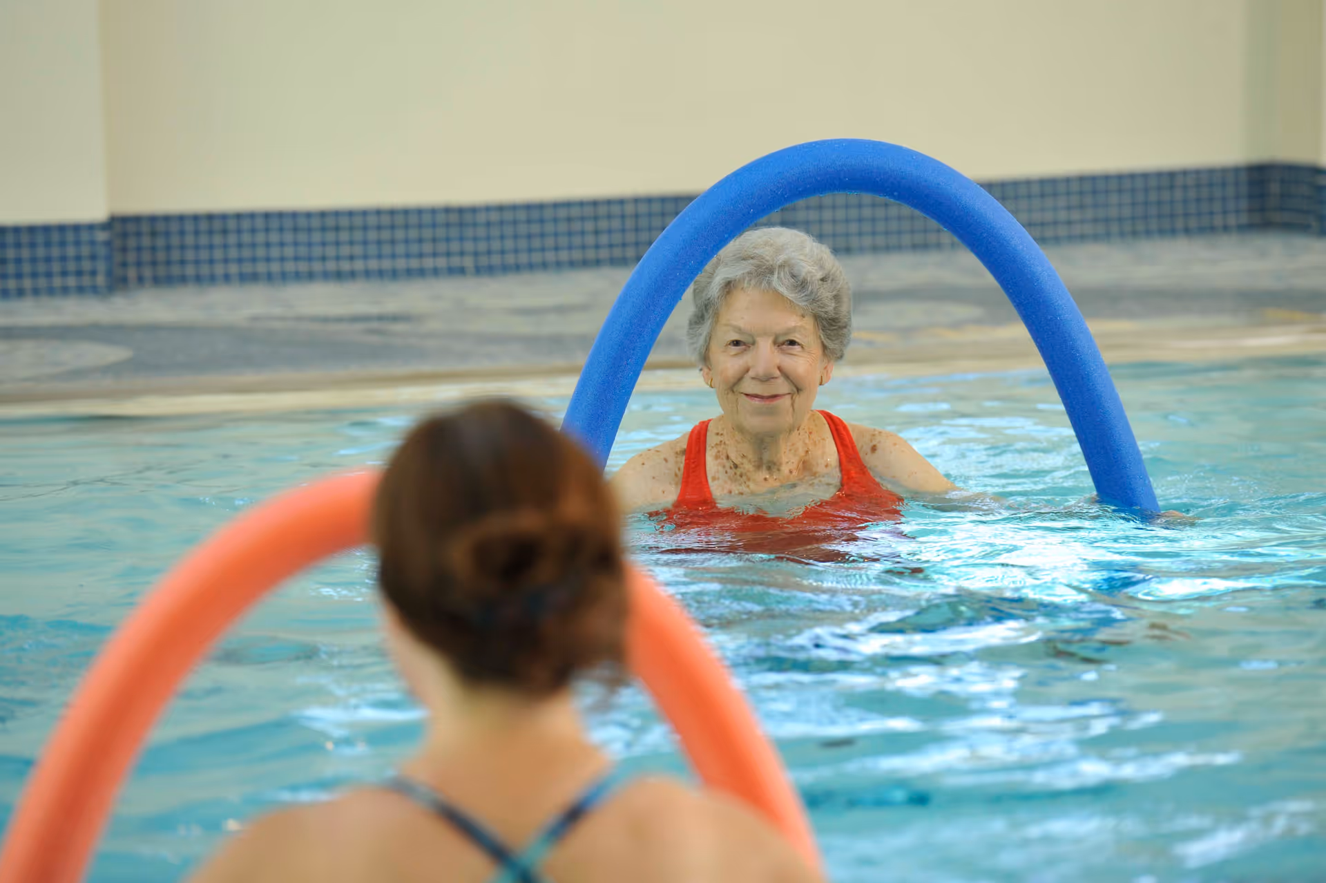 An elderly woman in a red swimsuit is in a swimming pool holding a blue pool noodle, facing a younger woman who is holding an orange pool noodle. The setting appears to be an indoor pool with tiled walls.